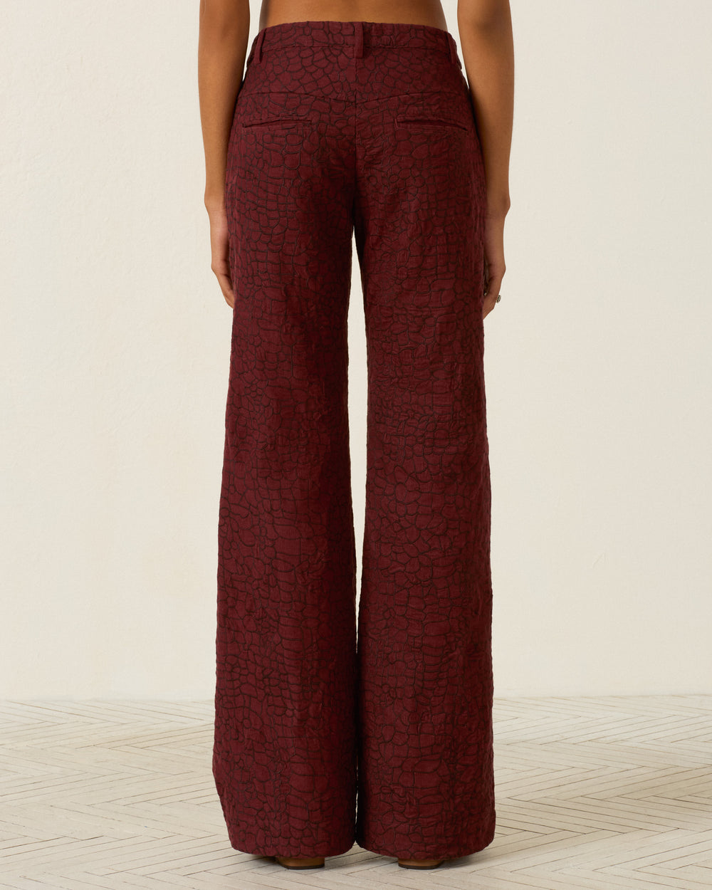 A person stands facing away, wearing the Cooper Auburn Crocodile Pant—high-waisted, wide-leg corduroy pants in a textured burgundy finish—on a light patterned floor with a plain background.
