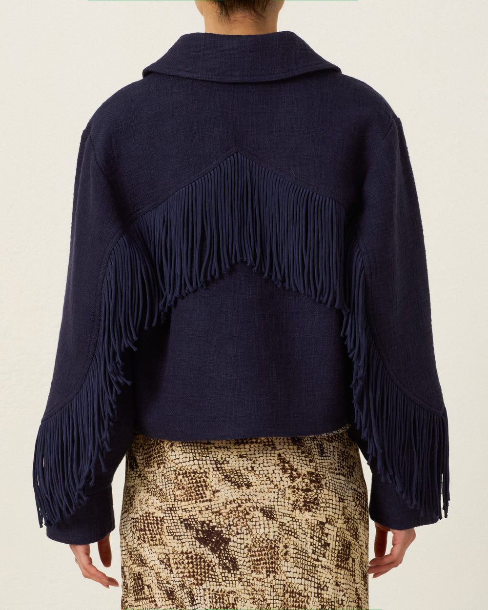 Person wearing the Court Indigo Night Jacket—a navy Khadi cotton cropped zip jacket featuring Western-style fringing in a V-shaped pattern on the back and sleeves—paired with a beige and brown snakeskin-patterned skirt.