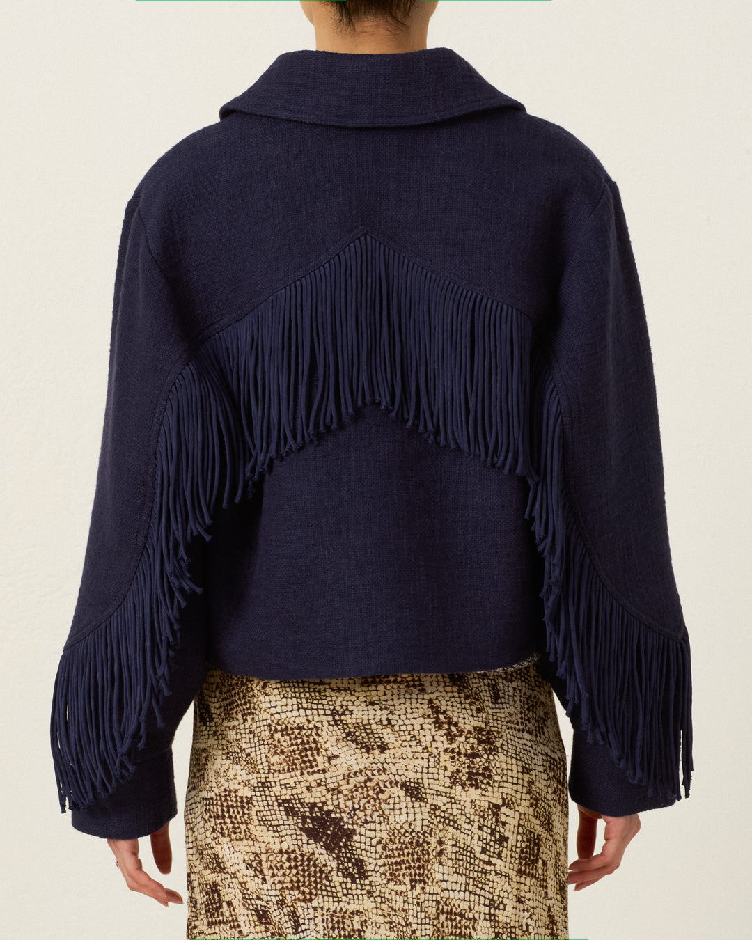 Person wearing the Court Indigo Night Jacket—a navy Khadi cotton cropped zip jacket featuring Western-style fringing in a V-shaped pattern on the back and sleeves—paired with a beige and brown snakeskin-patterned skirt.