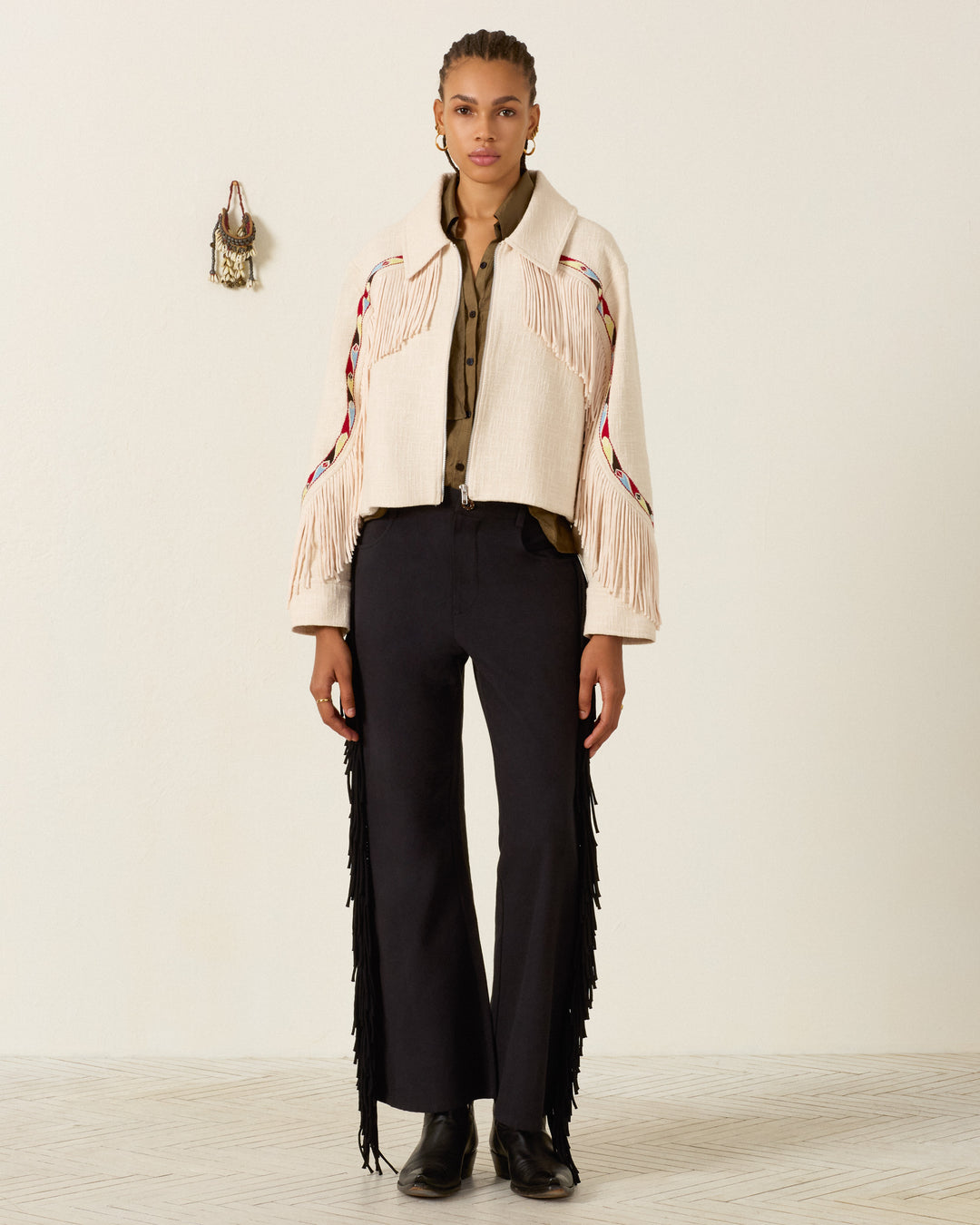 A person stands facing forward, wearing the cream Court Prairie Fringe Jacket made from Khadi cotton with Western-inspired fringing, an olive shirt, black fringed pants, and black boots, with a small decorative item hanging on the wall behind them.