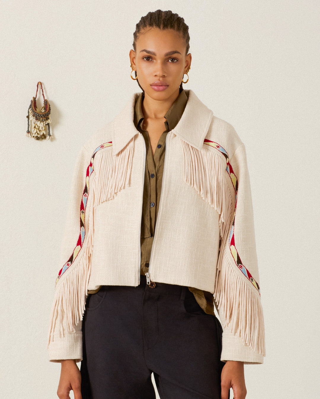 A woman wears the Court Prairie Fringe Jacket—a beige, Western-inspired design with patterned trim—over an olive shirt and black pants, standing against a plain background with wall decor visible to the left.