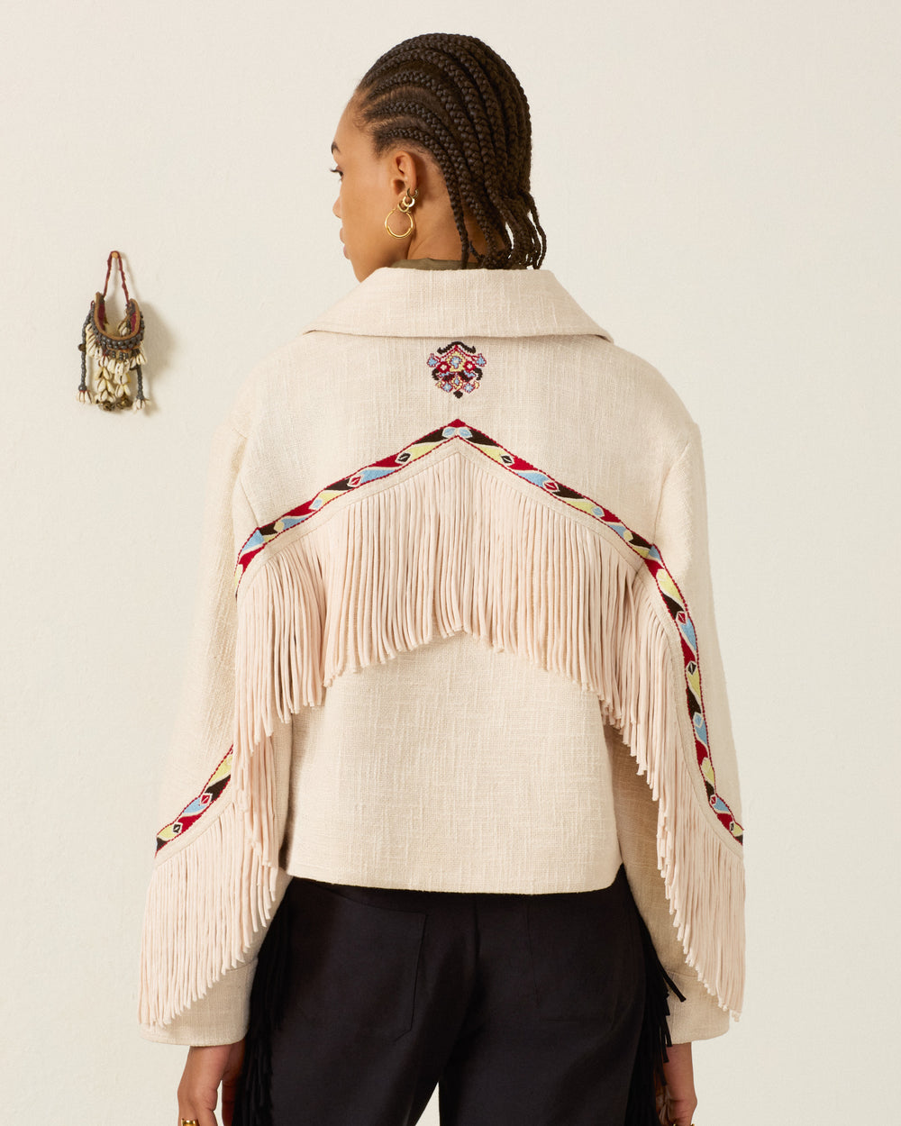 A person with braided hair wears the Court Prairie Fringe Jacket, a cream cropped zip-up style with Western-inspired fringing and colorful embroidered trim, paired with black pants and facing away from the camera against a plain wall.