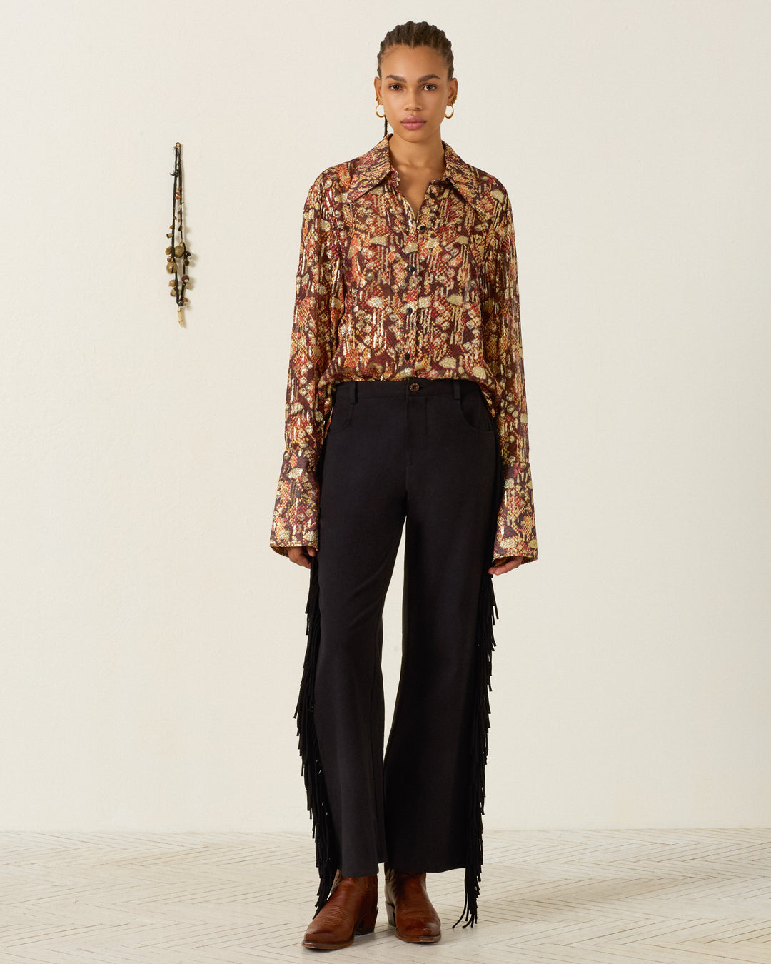 Person stands facing forward, wearing the Dana Topaz Snakeskin Shirt with a ’70s dagger collar, black pants featuring fringe on the sides, and brown boots; small wall decor appears in the background.