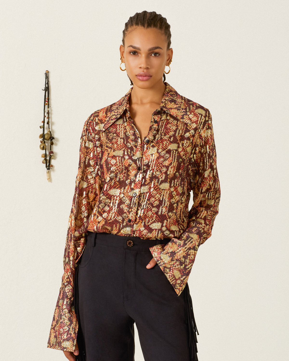 A woman with braided hair wears the Dana Topaz Snakeskin Shirt—a ’70s-inspired button-up with a dagger collar, patterned design, and flared sleeves—paired with black pants, standing against a plain wall adorned with a hanging string of beads.