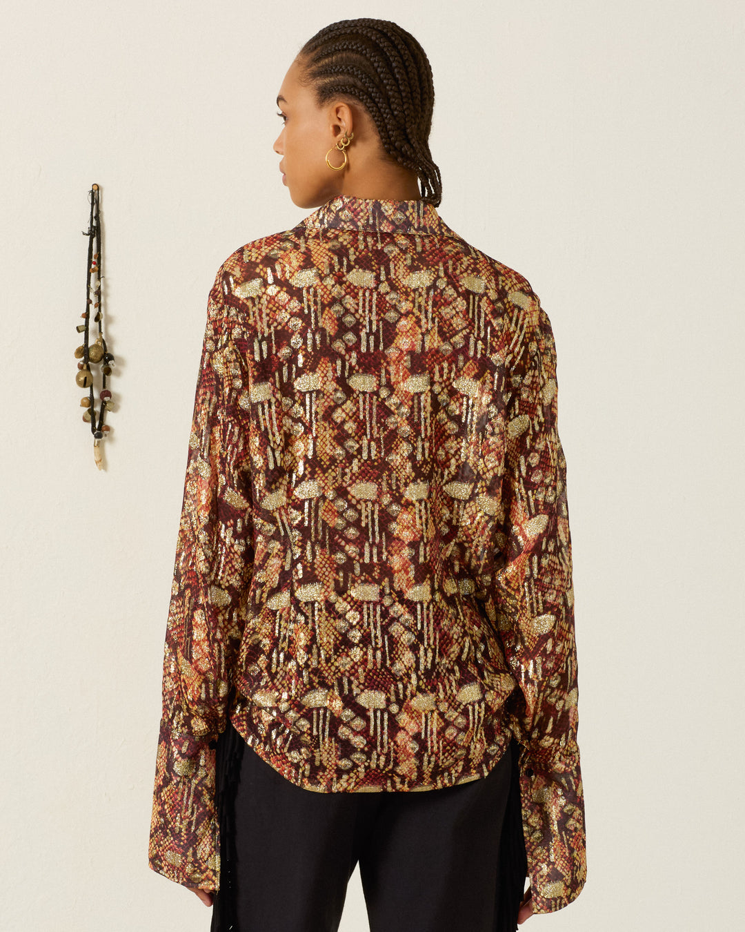 A person with braided hair wears the Dana Topaz Snakeskin Shirt, featuring long sleeves and a ’70s-inspired dagger collar, standing with their back to the camera. A decorative object hangs on the white wall to the left.