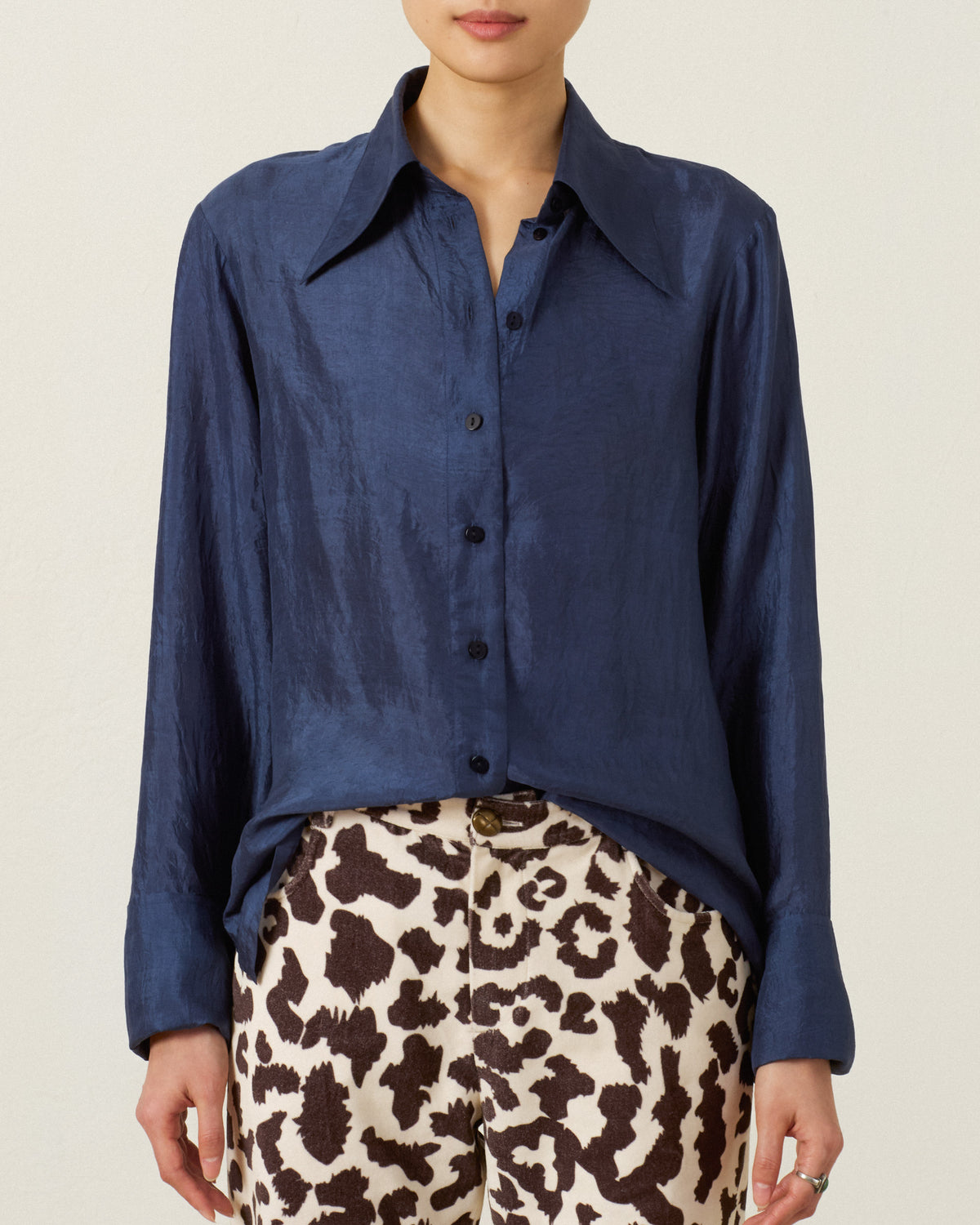 Wearing the Dana Indigo Night Silk Shirt—a dark blue, vintage-inspired piece featuring long sleeves, a wide collar, and button-up detailing—paired with black-and-white animal print pants, standing against a plain background.