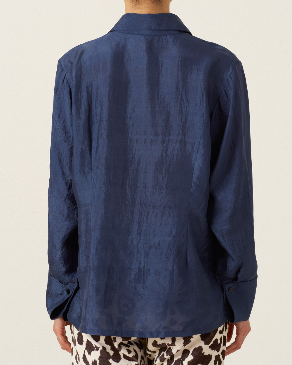 A person is seen from the back wearing the Dana Indigo Night Silk Shirt, a long-sleeve, navy blue hand-loomed silk button-up with a lustrous finish, paired with patterned brown and white pants.