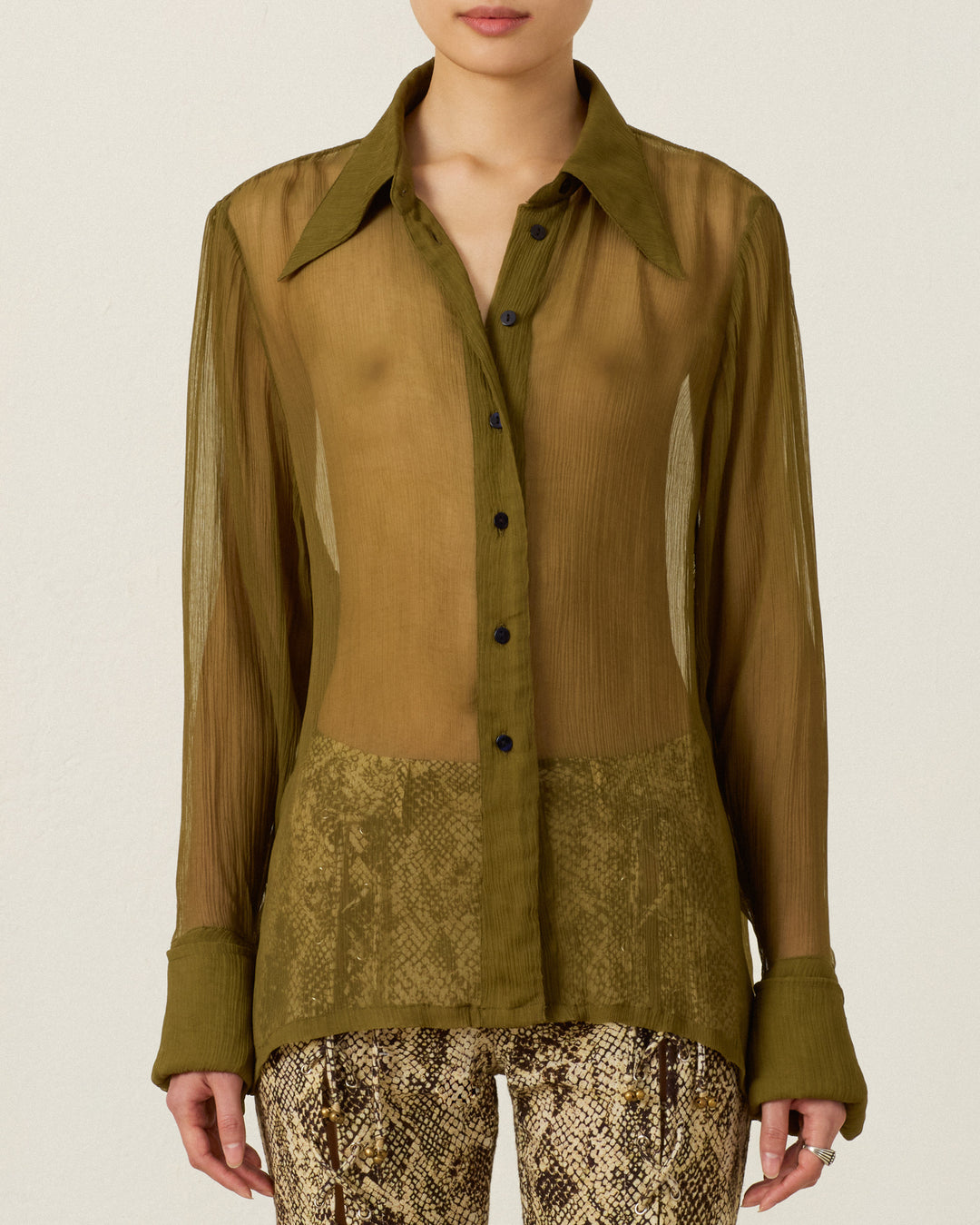 A person wearing the Dana Olive Chiffon Shirt—featuring sheer cupro-chiffon fabric, long sleeves with large cuffs, and a ’70s dagger collar—paired with brown snakeskin-patterned pants, stands against a plain background.