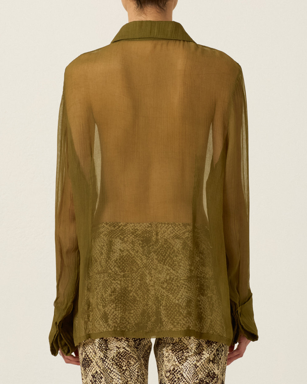 A person is seen from the back wearing the Dana Olive Chiffon Shirt, a sheer olive-green cupro-chiffon long-sleeve top with a ’70s dagger collar, paired with high-waisted, brown snake-print pants.