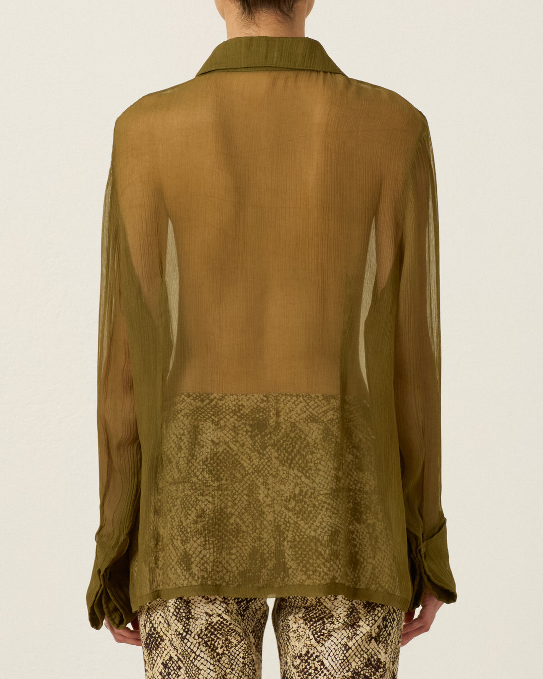 A person is seen from the back wearing the Dana Olive Chiffon Shirt, a sheer olive-green cupro-chiffon long-sleeve top with a ’70s dagger collar, paired with high-waisted, brown snake-print pants.