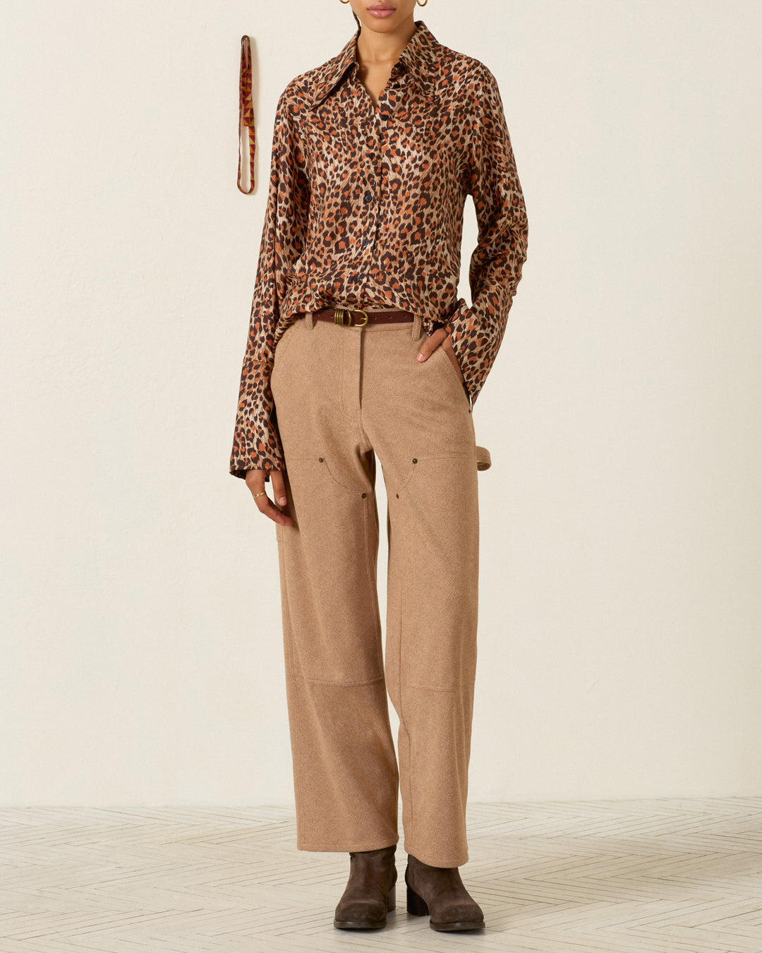 A person wearing a Dana Leopard Trek Silk Shirt, tan wide-leg trousers with a brown belt, and dark ankle boots.