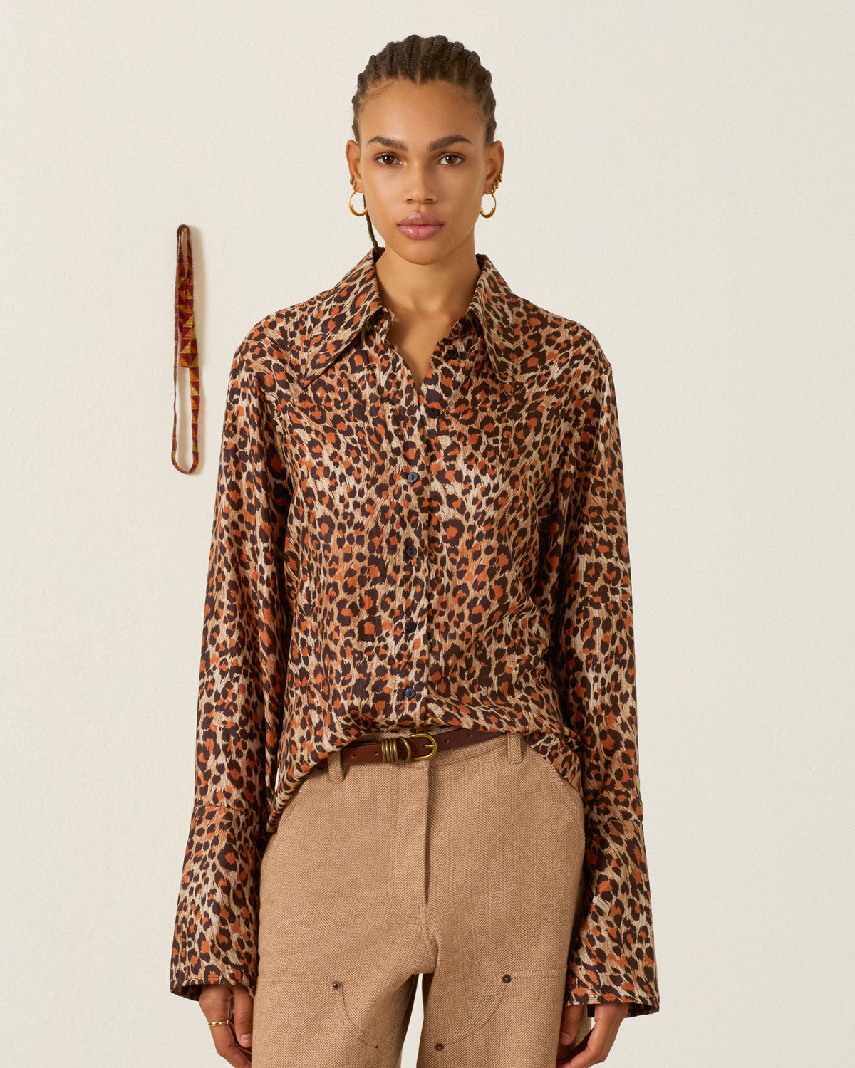 The Dana Leopard Trek Silk Shirt, a long-sleeved artisan top in a brown and black leopard print, is worn tucked into tan trousers with a brown belt.