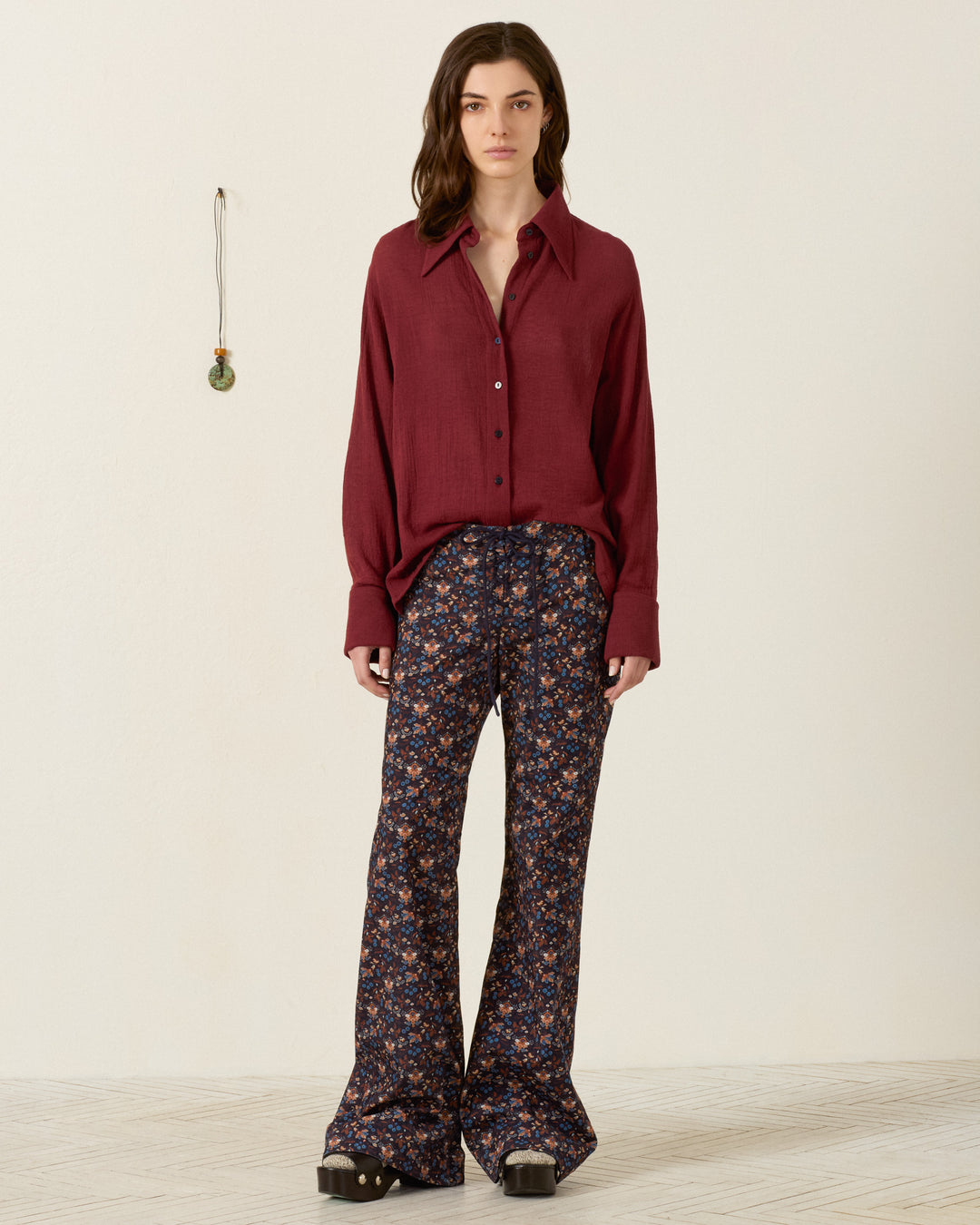 A person stands indoors against a neutral background with a small hanging decoration, wearing the Dana Mahogany Shirt—crafted from maroon cotton dobby with a subtle texture and featuring a ’70s dagger collar—paired with wide-leg floral pants and black platform shoes.