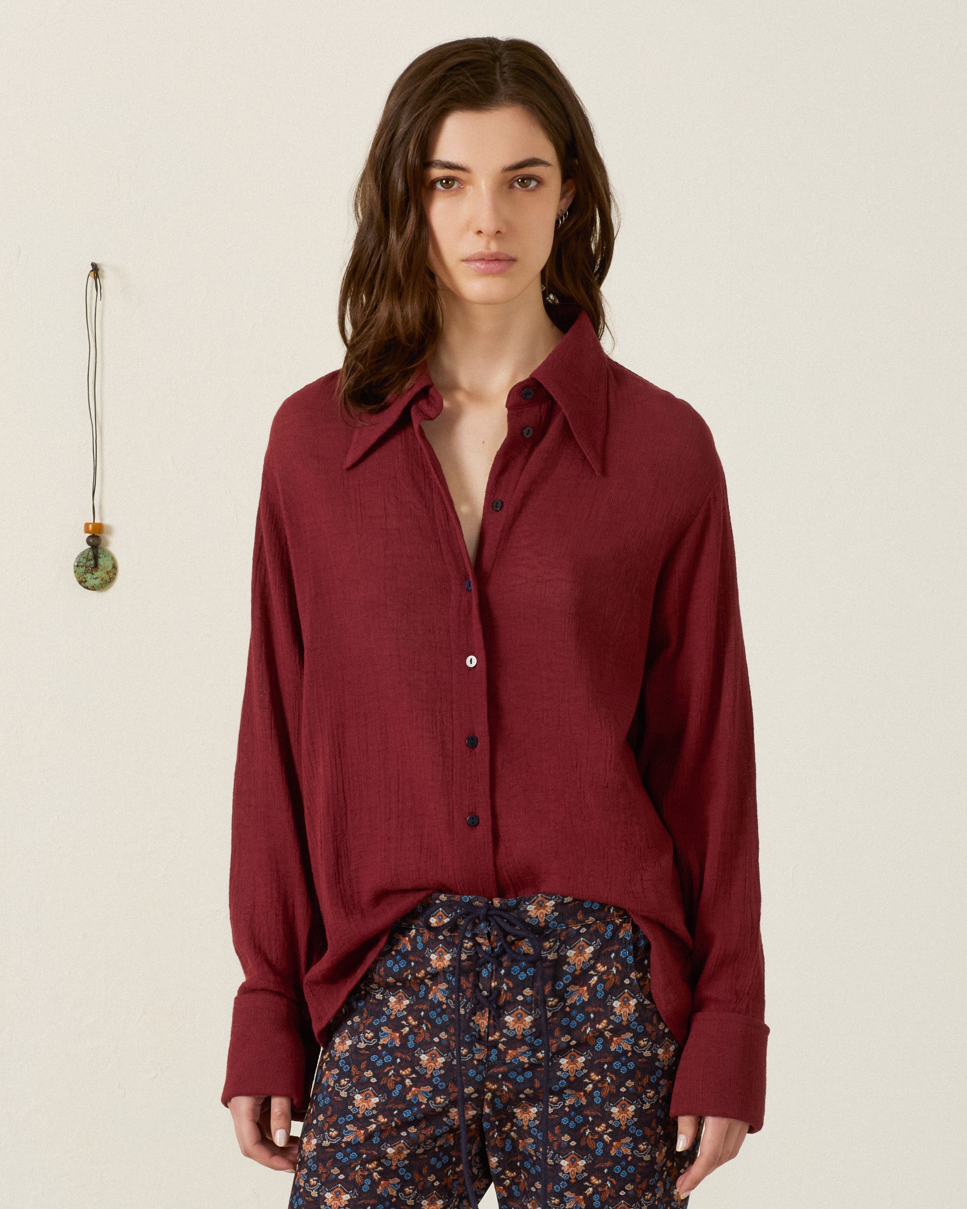 A person stands indoors wearing the Dana Mahogany Shirt, a loose, vintage-inspired burgundy button-up with a ’70s dagger collar, paired with floral drawstring pants, as a pendant hangs on the wall nearby.