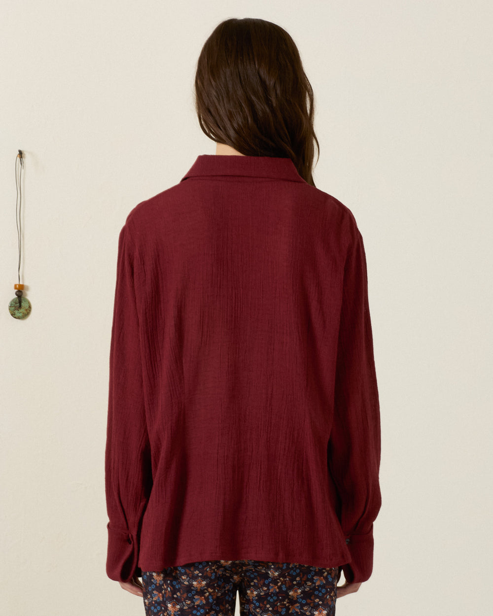 A person with long brown hair stands facing away, wearing the Dana Mahogany Shirt—a vintage-inspired maroon piece with a ’70s dagger collar—paired with floral patterned pants, set against a plain wall with a hanging decoration on the left.