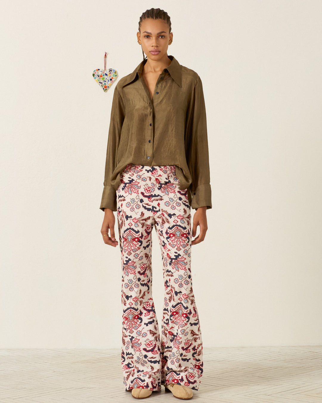 A person stands against a plain wall, wearing the Dana Olive Branch Silk Shirt with French cuffs and wide-leg, patterned pants. A small decorative heart hangs on the wall beside them.