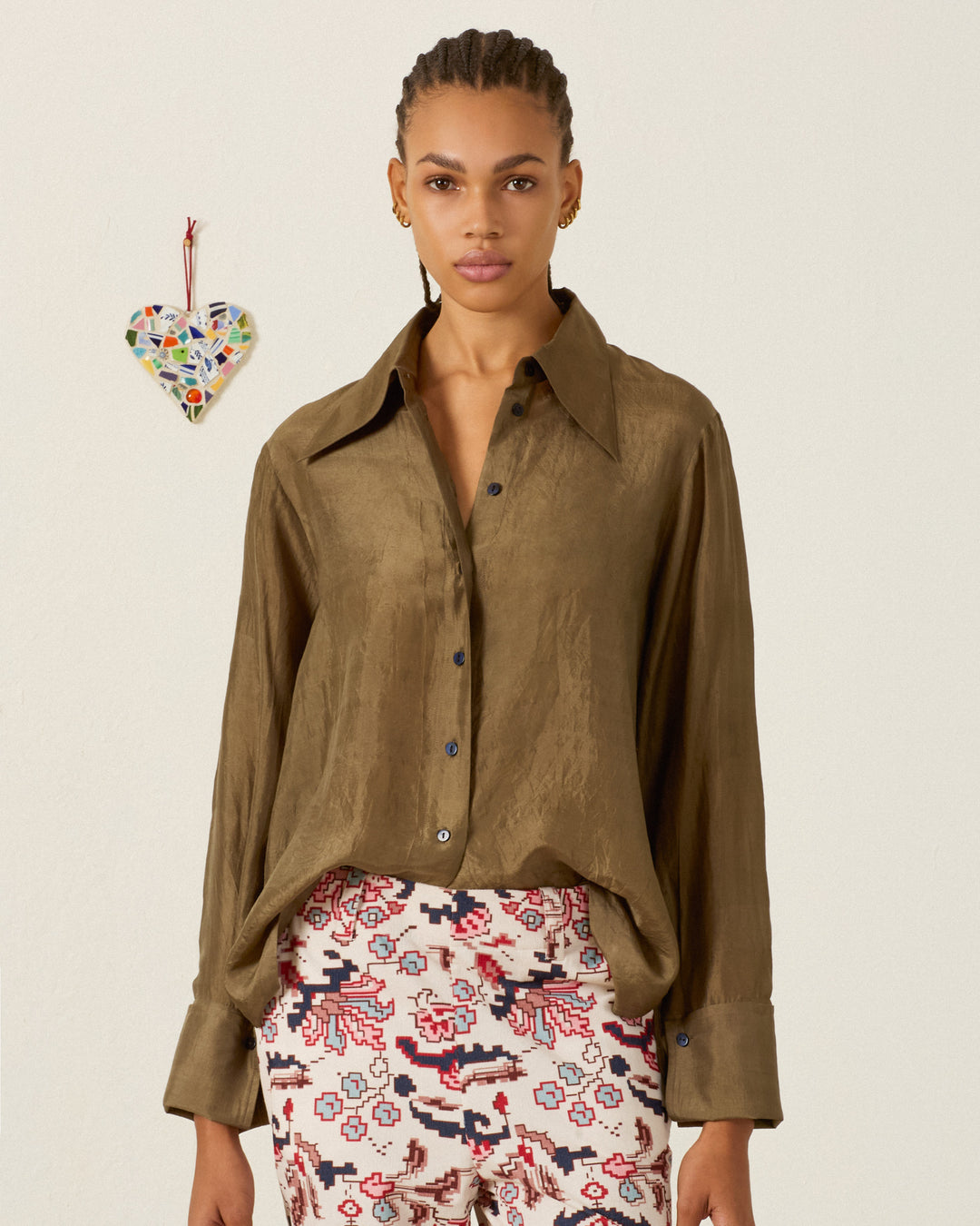 A woman wearing the Dana Olive Branch Silk Shirt with French cuffs and patterned pants stands against a plain background adorned with a mosaic heart decoration on the wall.