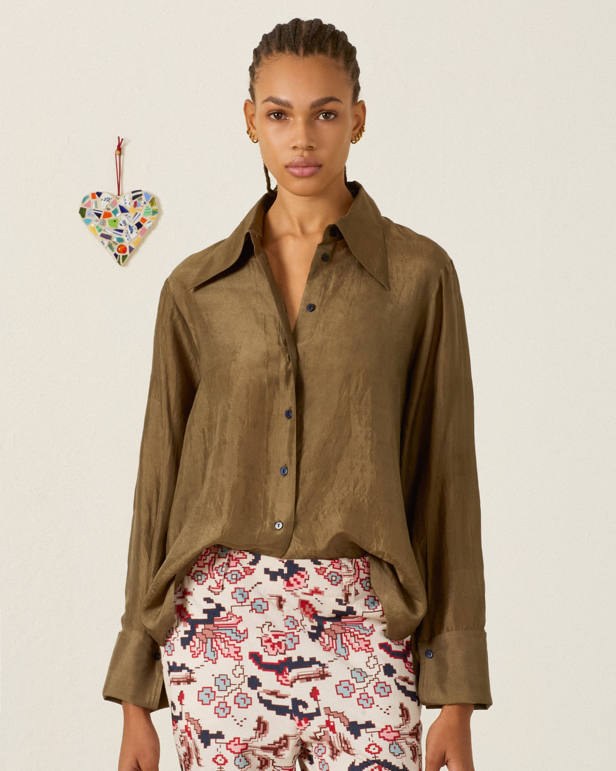 A woman wearing the Dana Olive Branch Silk Shirt with French cuffs and patterned pants stands against a plain background adorned with a mosaic heart decoration on the wall.
