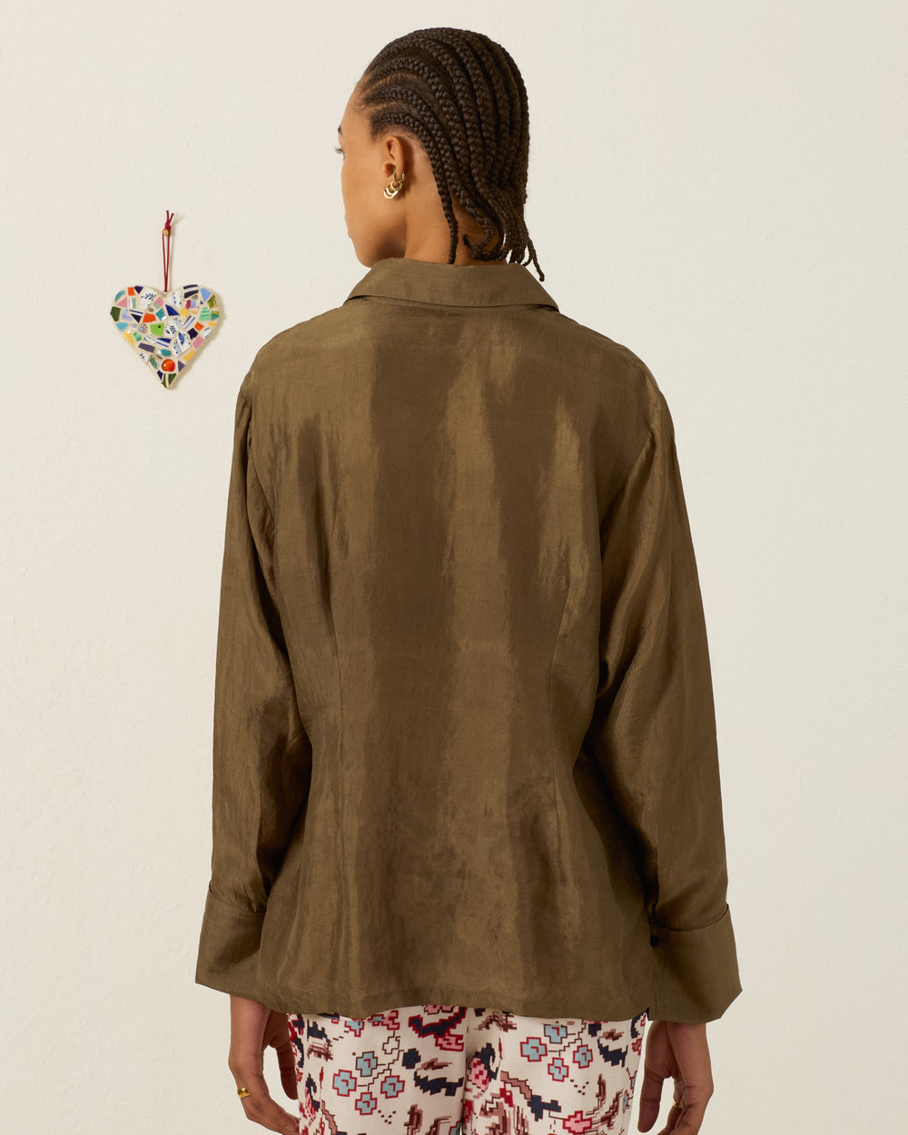 A person with braided hair, wearing the Dana Olive Branch Silk Shirt—an olive green, vintage-inspired top with French cuffs—and patterned pants stands facing a wall adorned with a small mosaic heart decoration.