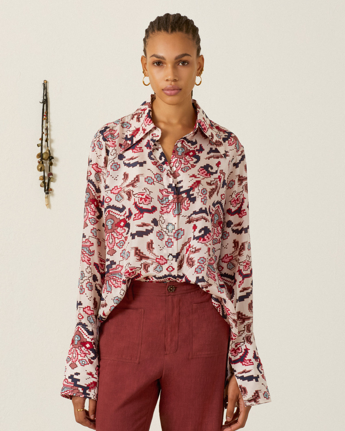 A woman stands against a plain wall, wearing the Dana Horizon Kilim Silk Shirt—hand-loomed silk adorned with red and blue kilim-inspired motifs—paired with high-waisted red pants. Jewelry hangs on the wall in the background.