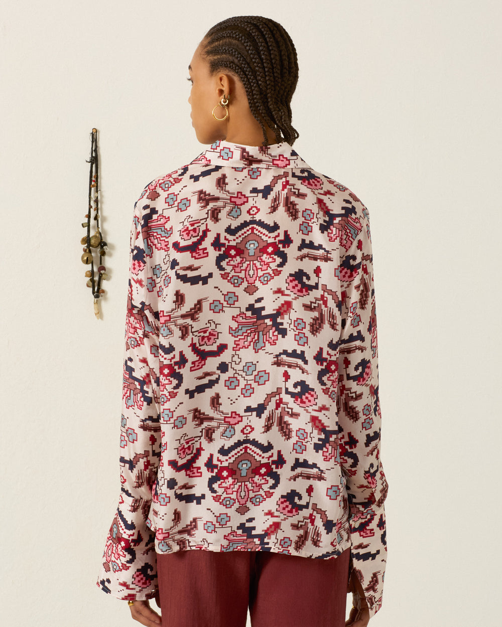 A person with braided hair faces away, wearing the Dana Horizon Kilim Silk Shirt—a long-sleeve, hand-loomed silk shirt featuring a red, black, and pink abstract pattern inspired by kilim motifs. A beaded wall hanging decorates the light-colored wall in the background.