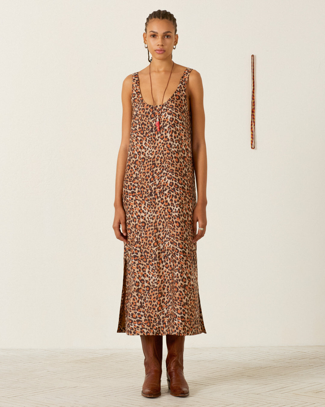 Wearing the Daph Leopard Trek Silk Dress and brown boots, the person stands against a plain white wall, with a vertical patterned stick displayed behind them.