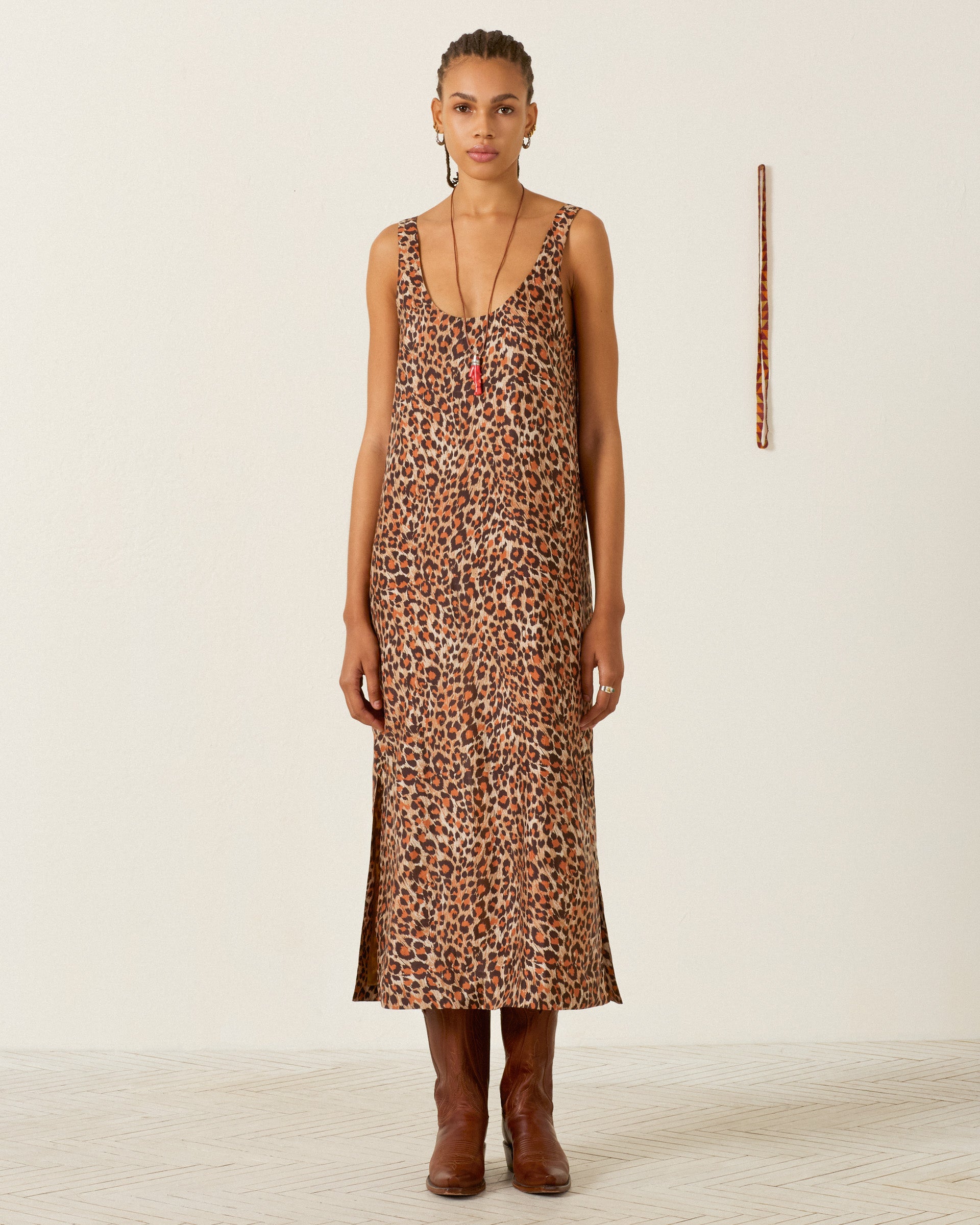 Wearing the Daph Leopard Trek Silk Dress and brown boots, the person stands against a plain white wall, with a vertical patterned stick displayed behind them.