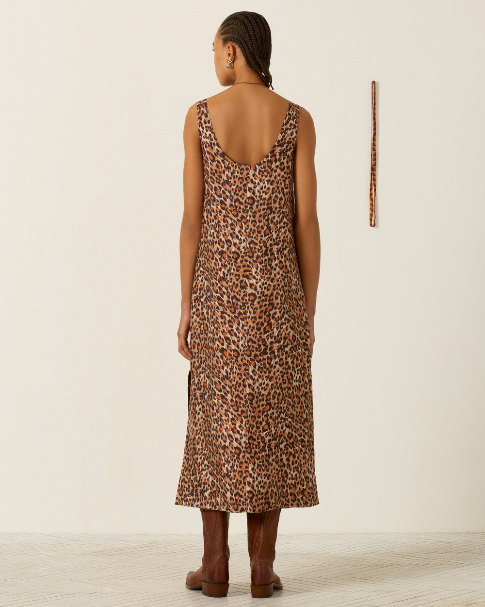 A person with braided hair stands facing away, wearing the Daph Leopard Trek Silk Dress—an artisan, sleeveless leopard-print piece—paired with brown boots in a minimalist room featuring a vertical wooden stick on the wall.
