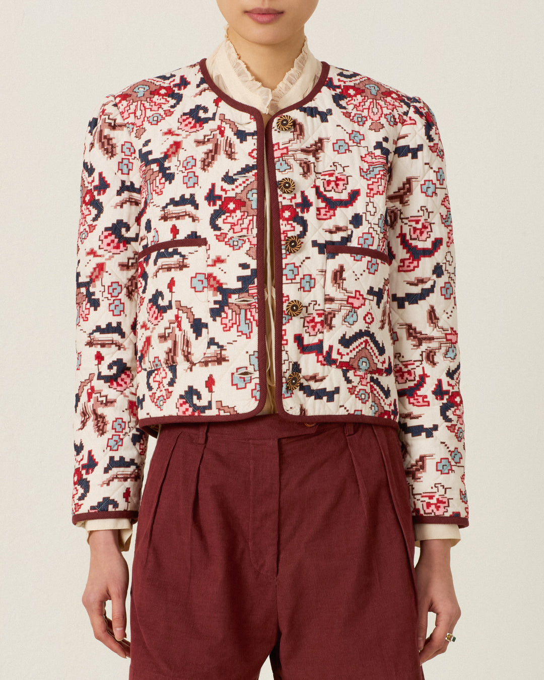 Person wearing the Dorian Horizon Kilim Jacket—a patterned, cream and red collarless quilted jacket—paired with a high-neck blouse and maroon pleated pants, standing against a plain background.