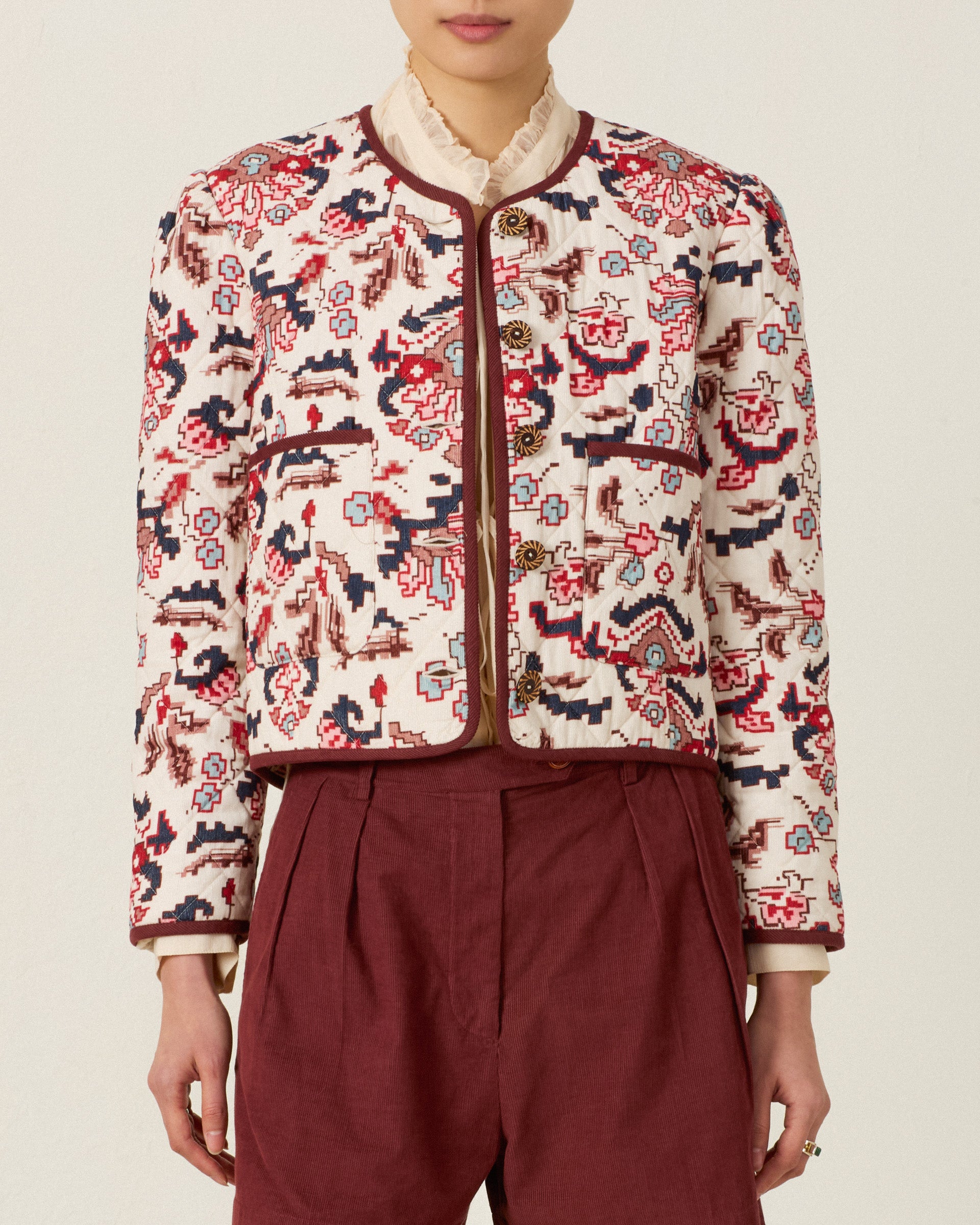Person wearing the Dorian Horizon Kilim Jacket—a patterned, cream and red collarless quilted jacket—paired with a high-neck blouse and maroon pleated pants, standing against a plain background.
