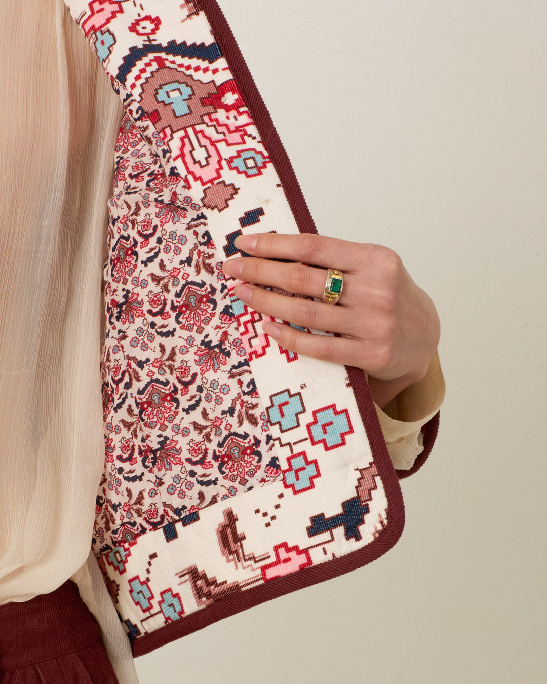 Wearing the Dorian Horizon Kilim Jacket—distinguished by its patterned, collarless design and cropped silhouette—a person opens it to reveal the jacket’s striking floral lining. They pair it with a sheer, cream-colored blouse and a green stone ring.
