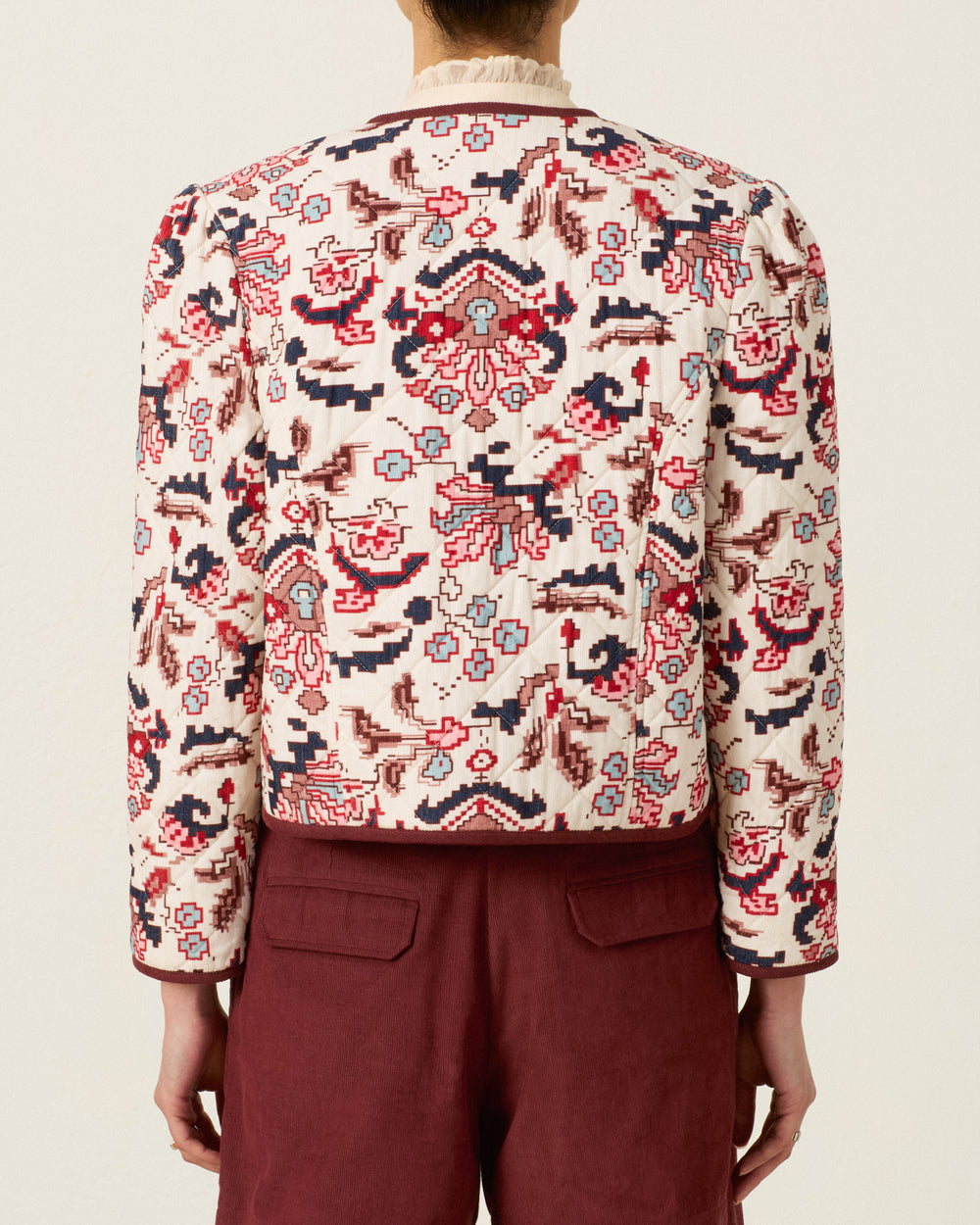 A person is shown from the back against a plain background, wearing the Dorian Horizon Kilim Jacket—a long-sleeve, collarless piece featuring red and blue patterns and a cropped silhouette—paired with maroon corduroy trousers.