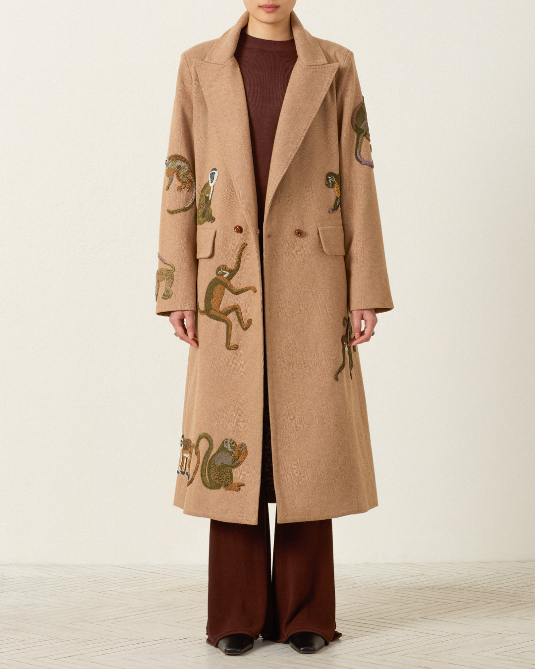 A person wearing the Duke Barrel of Monkeys Coat—a long beige wool herringbone topcoat featuring embroidered monkeys and handcrafted nautilus shell buttons—over a brown top and pants, stands against a plain light background.