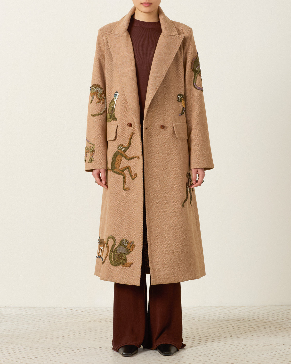 A person wearing the Duke Barrel of Monkeys Coat—a long beige wool herringbone topcoat featuring embroidered monkeys and handcrafted nautilus shell buttons—over a brown top and pants, stands against a plain light background.