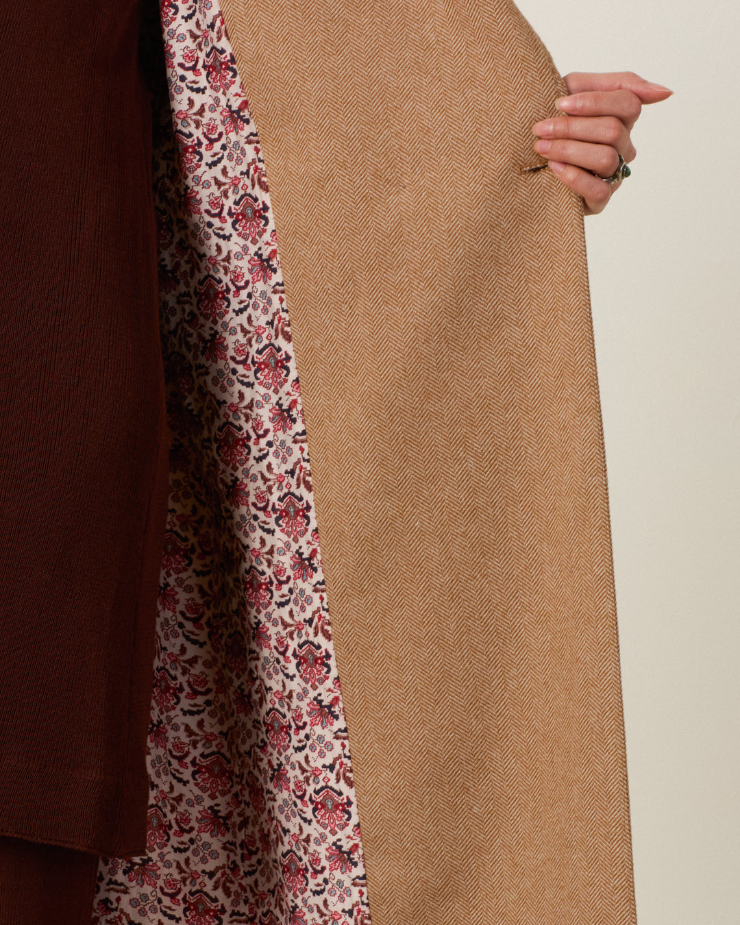 A hand pulls open the Duke Barrel of Monkeys Coat, crafted from tan wool herringbone, to reveal its floral patterned lining alongside a section of dark brown fabric.