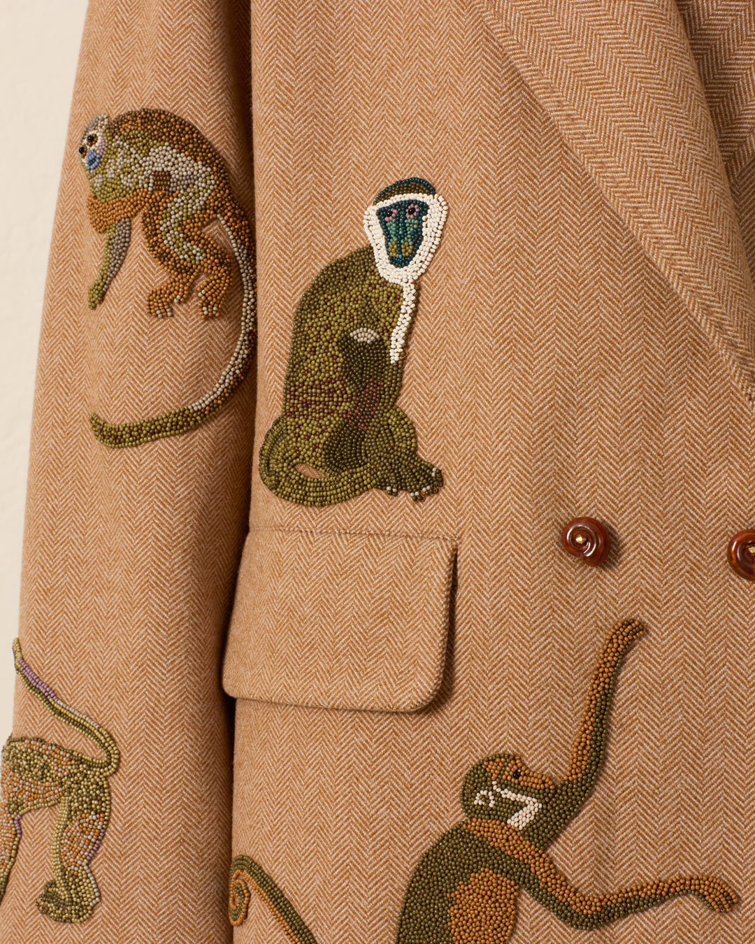 Introducing the Duke Barrel of Monkeys Coat: a tan wool herringbone blazer adorned with intricately embroidered and hand-beaded monkeys in various poses around the pocket and lapel. This distinctive piece is finished with handcrafted nautilus shell buttons.