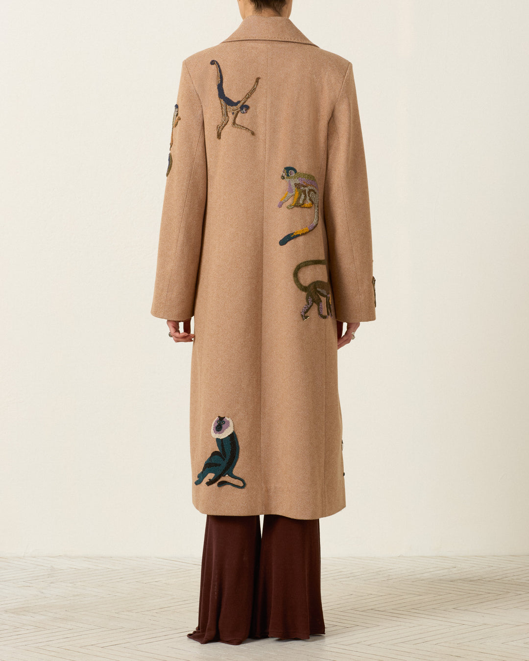 A person is standing indoors on a light patterned floor, facing away from the camera, wearing the Duke Barrel of Monkeys Coat—a long tan wool herringbone topcoat featuring embroidered monkeys on the back and handcrafted nautilus shell buttons.