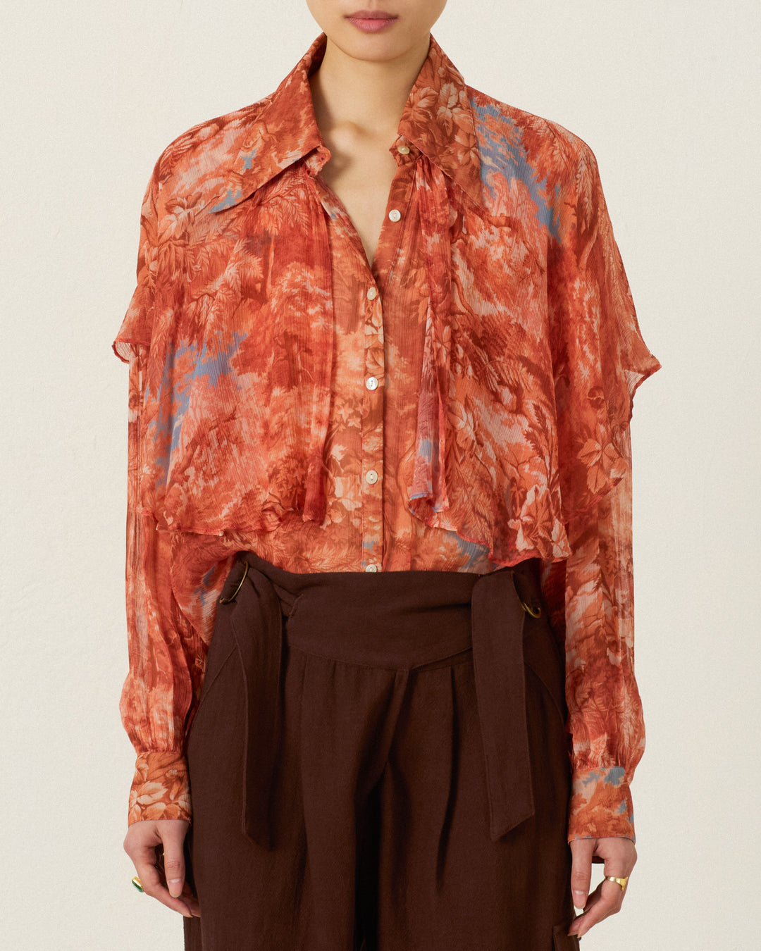 A woman wears the Eleanor Heritage Canopy Shirt—a rust-orange floral cape blouse with a tie detail at the neck and long sleeves—paired with high-waisted brown trousers.