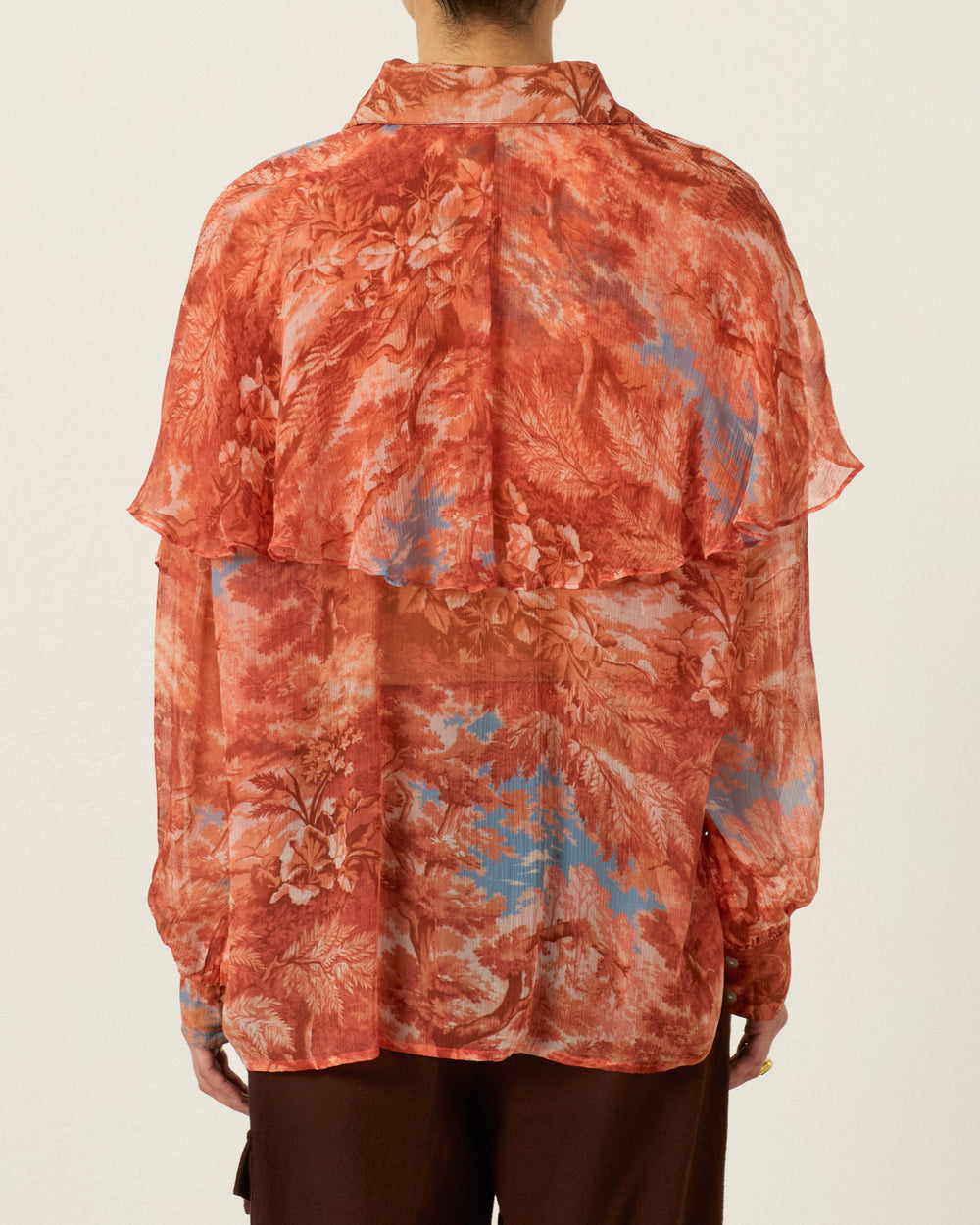 A person is seen from behind against a plain background, wearing the Eleanor Heritage Canopy Shirt—a long-sleeve, sheer red floral cupro-chiffon cape blouse featuring a layered back detail and a subtle ’70s dagger collar.