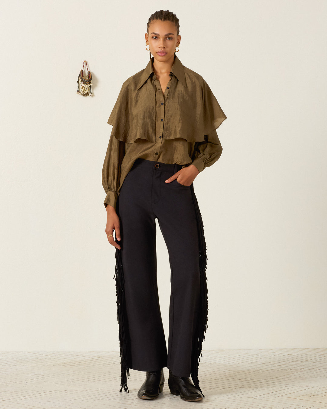 A person standing indoors wears a tan button-up shirt with layered cape detail, the Bunny Onyx Fringe Pant—black Western-inspired wide-leg pants with fringe sides—and black shoes. A small decorative item hangs on the wall behind them.