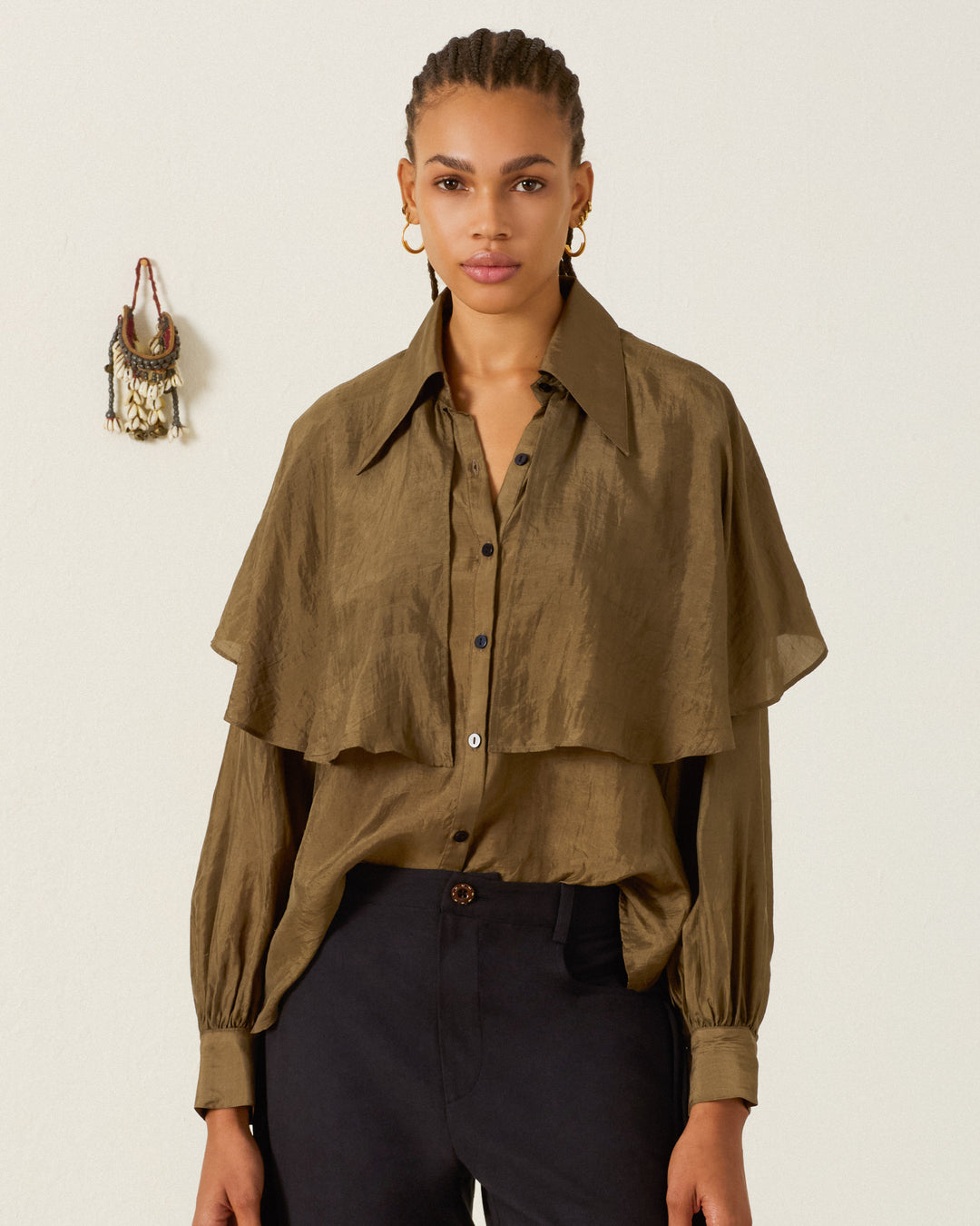 A woman stands against a plain light-colored wall with a small hanging ornament in the background, wearing the Eleanor Olive Silk Shirt—an olive green, hand-loomed silk blouse featuring a subtle ’70s dagger collar—and black pants.