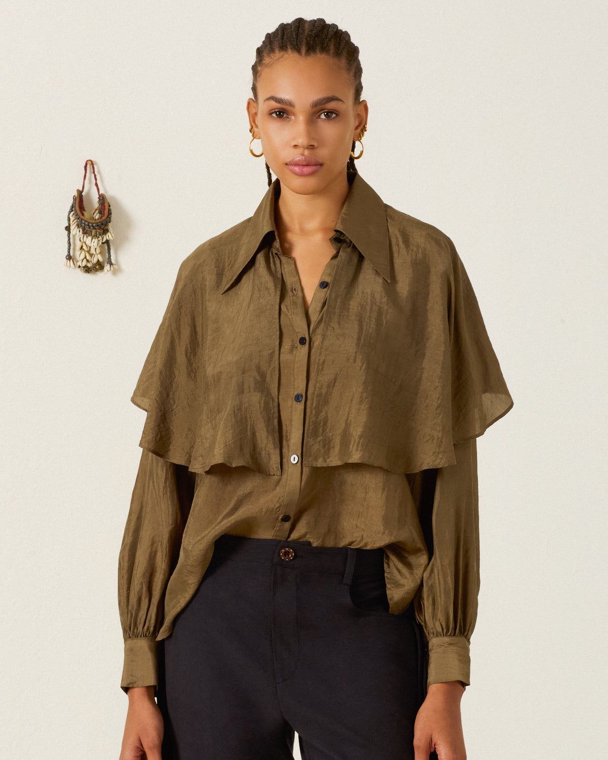 A woman stands against a plain light-colored wall with a small hanging ornament in the background, wearing the Eleanor Olive Silk Shirt—an olive green, hand-loomed silk blouse featuring a subtle ’70s dagger collar—and black pants.