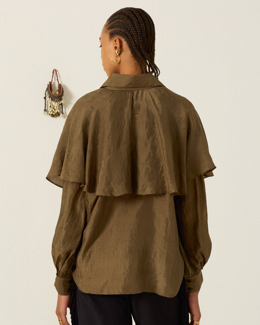 A person with braided hair, dressed in the Eleanor Olive Silk Shirt—a brown, long-sleeved hand-loomed silk blouse featuring a ruffled cape detail—stands facing away from the camera, with a small wall decoration visible on a plain light background.