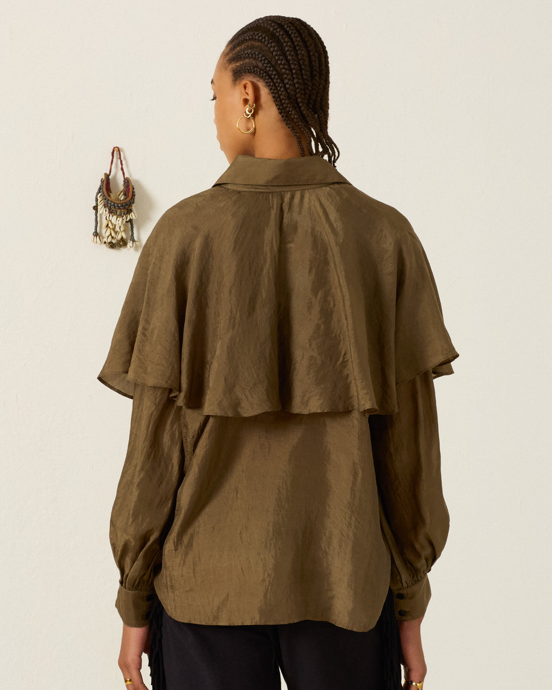 A person with braided hair, dressed in the Eleanor Olive Silk Shirt—a brown, long-sleeved hand-loomed silk blouse featuring a ruffled cape detail—stands facing away from the camera, with a small wall decoration visible on a plain light background.