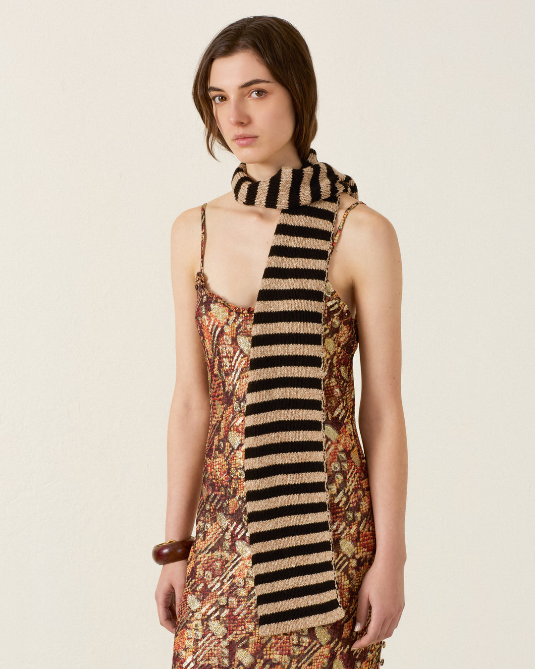 A person wearing a patterned sleeveless dress and the Elowen Zebra Stripe Scarf, featuring cozy black and beige stripes, stands against a plain background.
