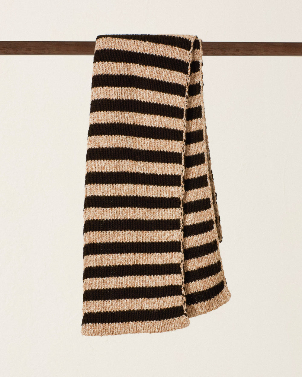 The Elowen Zebra Stripe Scarf, featuring cozy horizontal black and beige bands, is draped over a dark wooden rod against a plain light-colored background.