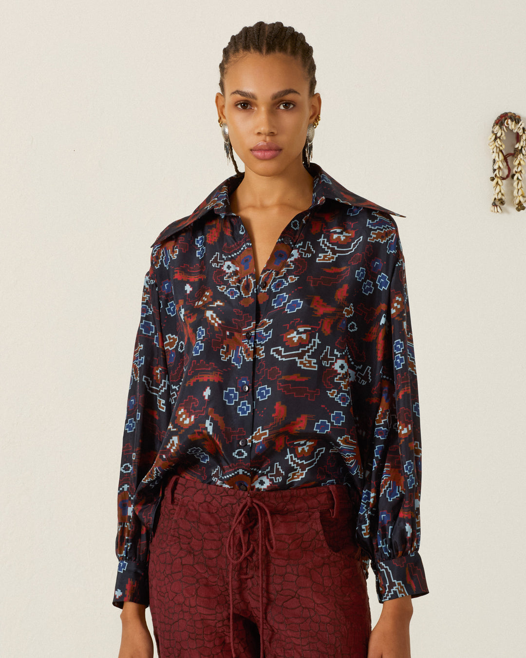 A woman stands against a light wall, wearing the Emery Indigo Kilim Silk Shirt—an artisan blouse featuring kilim-inspired motifs and a large collar—paired with red textured pants, as she looks directly at the camera.