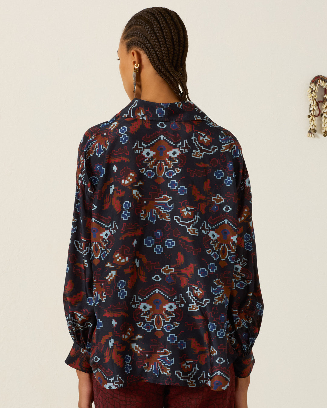 A person with braided hair, wearing the Emery Indigo Kilim Silk Shirt—a dark, patterned long-sleeve artisan blouse featuring kilim-inspired motifs—stands with their back to the camera against a plain light-colored wall.