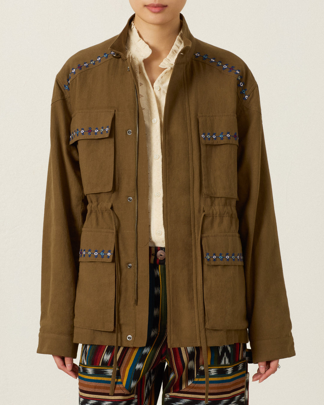 Evelyn Olive Branch Jacket in olive green Khadi cotton with four front pockets, a drawstring waist, and blue and white hand-embroidered trim along the shoulders and pockets.