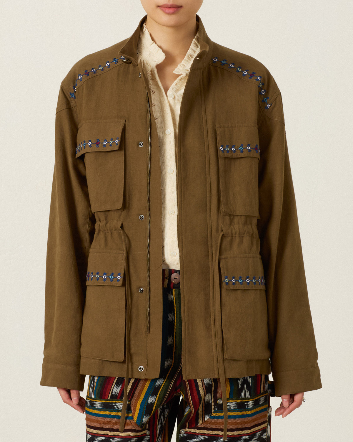 Evelyn Olive Branch Jacket in olive green Khadi cotton with four front pockets, a drawstring waist, and blue and white hand-embroidered trim along the shoulders and pockets.